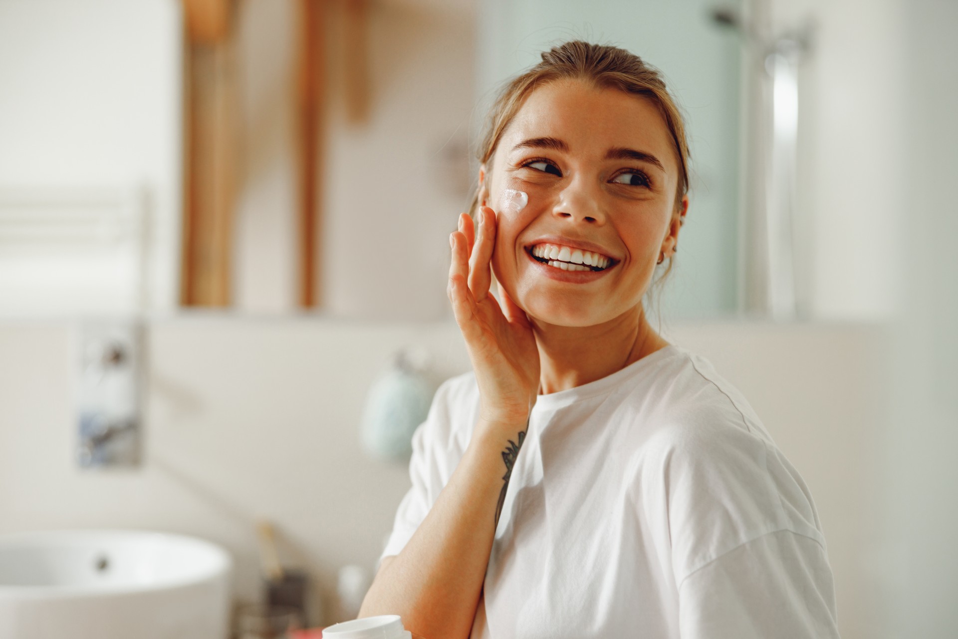 Young beautiful woman taking care of skin by applying moisturizer cream in bathroom